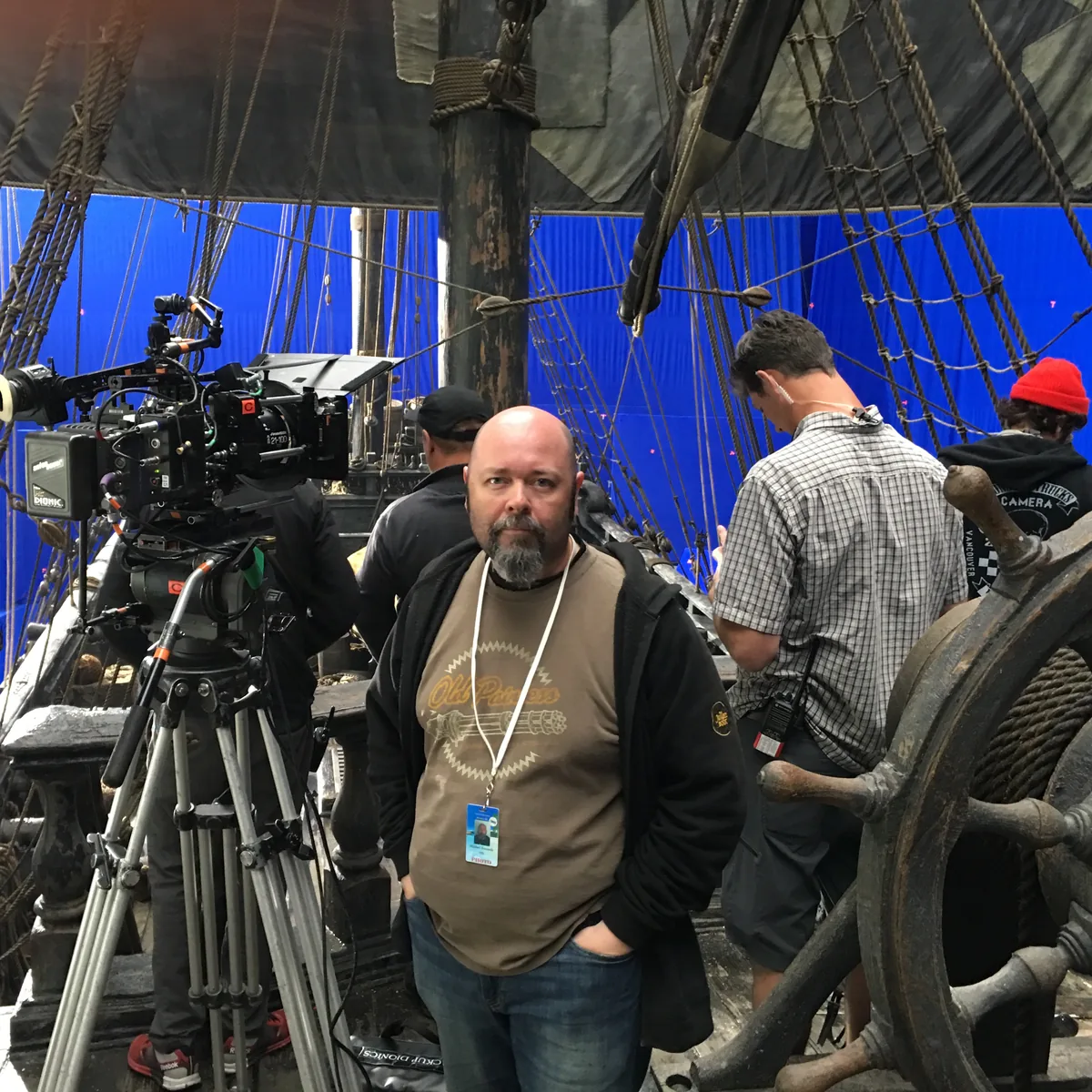 Michael Kennedy on the deck of a pirate ship set, standing beside a cinema camera and the ship's wheel, with a blue screen behind.
